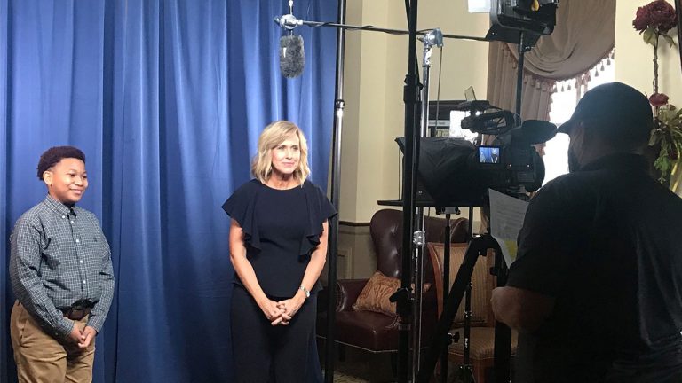 Hosts of Junior Achievement Business Hall of Fame standing in front of a blue curtain with a camera pointed at them