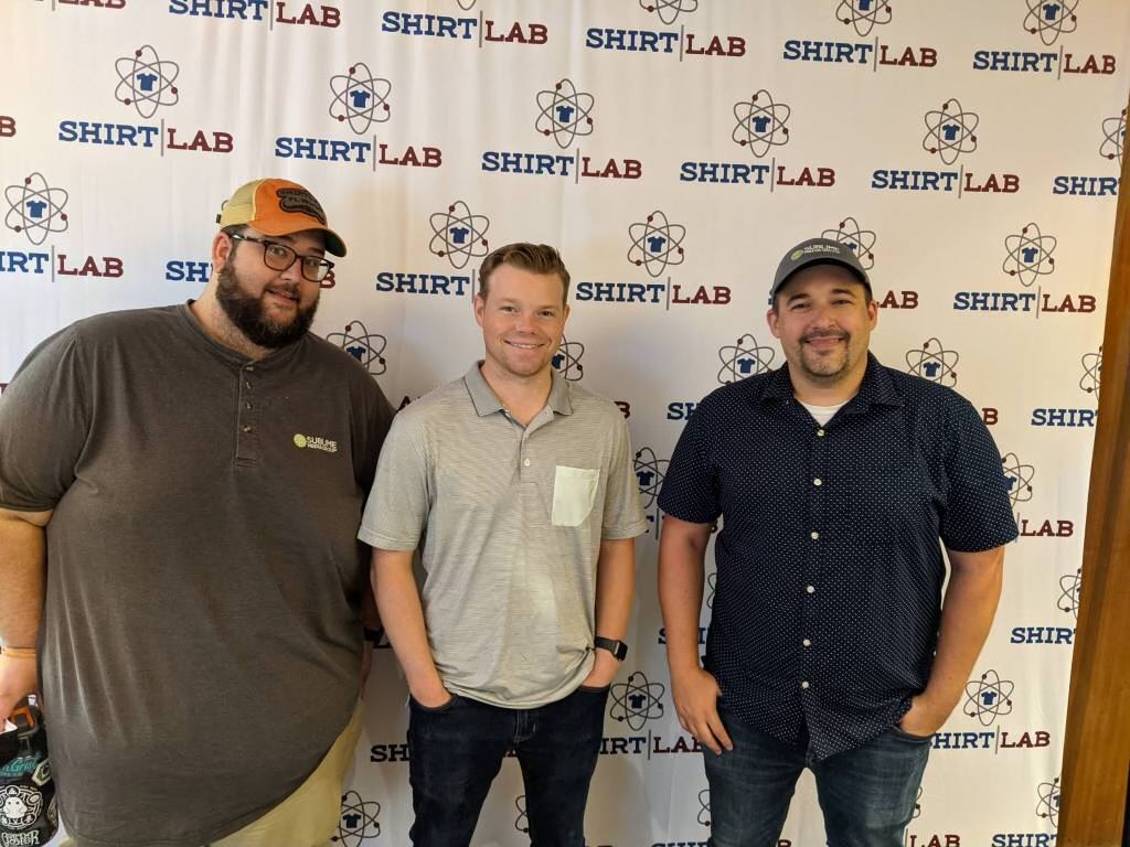 Will Kronenberger, Nick Johnson and Austin Albany standing in front of a step and repeat wall with the Shirt Lab logo