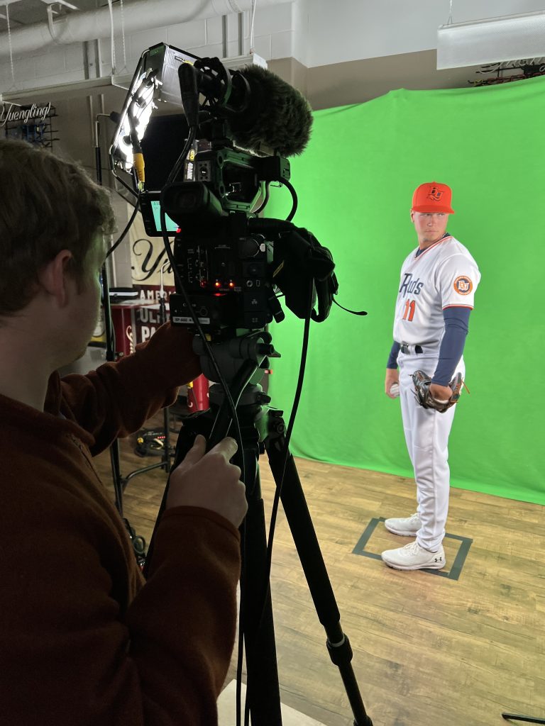 A baseball player stands in front of a green screen. A man with a camera is visible in the left side of the photo.