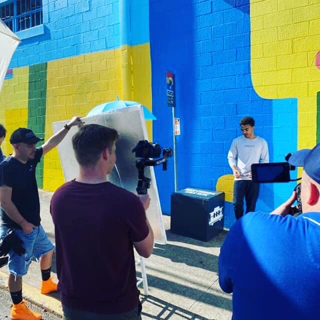 behind the scenes photo of a video shoot in front of a blue wall with a young male modeling a sweatshirt showing how you share company product videos to attract top talent