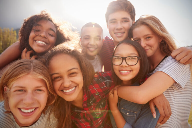 A diverse group of young people huddle together and smile while looking at the viewer. This image contains a lot of diversity.