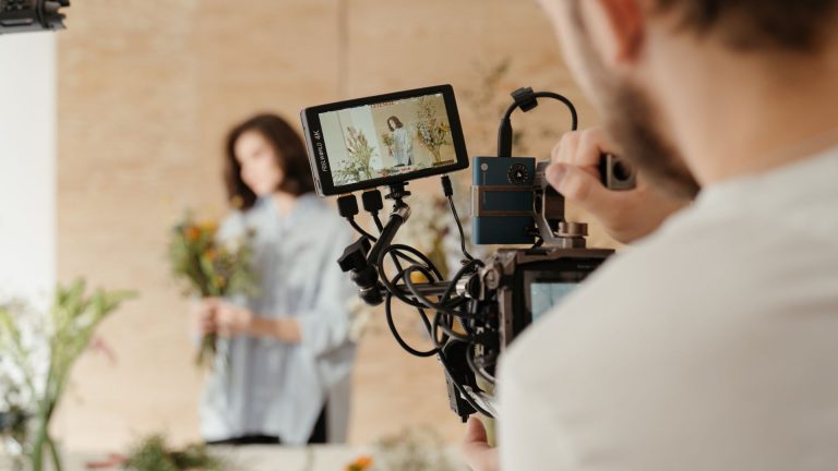 Photo of a man operating a camera to create videos of a woman building a floral bouquet.